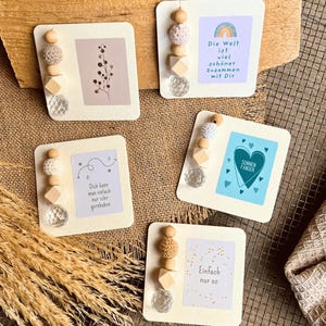 May include: Five square cards featuring decorative bead strands attached to the left side. Each card displays unique illustrations or German text, including phrases like Die Welt ist viel schöner zusammen mit Dir, Dich kann man einfach nur sehr gernhaben, Sonnen Fanger, and Einfach nur so.