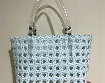 Puede incluir: Un bolso tote tejido con un patrón blanco y verde. El bolso tiene asas de plástico transparente sujetas con remaches plateados. El bolso está sobre una superficie rosa. El diseño tejido del bolso crea un patrón similar a una cuadrícula.