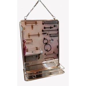 Op de afbeelding: Een rechthoekige spiegel met een gouden frame en een klein plankje, opgehangen aan een ketting. De spiegel reflecteert een badkamer met armaturen zoals handdoekrekken en een wastafel. Het ontwerp is vintage-geïnspireerd.
