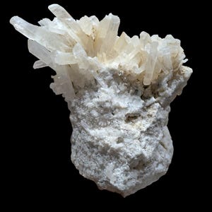 May include: A cluster of translucent, off-white crystal formations atop a rough, textured, white rock base. The crystals are pointed and clustered, with a natural, organic appearance against a black background.