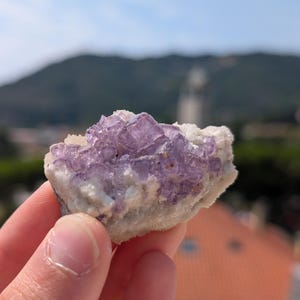 May include: A hand holds a natural fluorite crystal cluster. The crystal features a mix of translucent purple and white colors. The background shows a blurred view of a mountain and buildings under a blue sky.