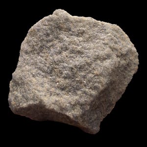 May include: A close-up of a rough, gray rock against a black background. The rock has a textured surface with small, sparkling particles. The overall color is a mix of gray and tan, with a slightly irregular shape. The rock appears to be a natural stone.