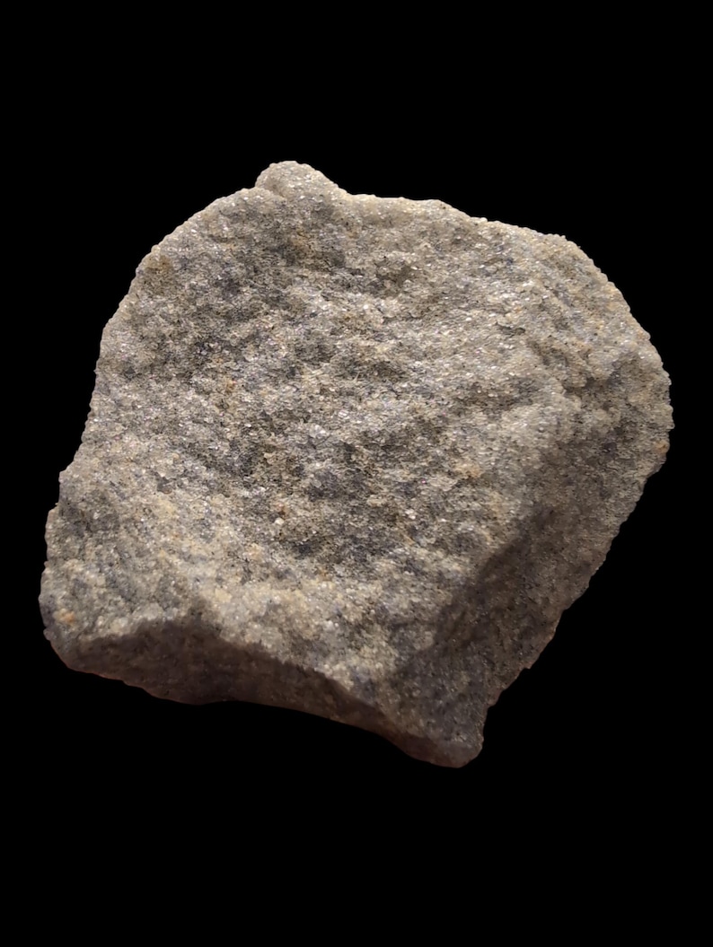 May include: A close-up of a rough, gray rock against a black background. The rock has a textured surface with small, sparkling particles. The overall color is a mix of gray and tan, with a slightly irregular shape. The rock appears to be a natural stone.
