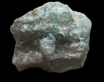 May include: A rough, uncut, light green mineral specimen against a black background. The crystalline structure of the stone is visible, with areas of translucence and a slightly rough texture. The overall shape is irregular and natural.