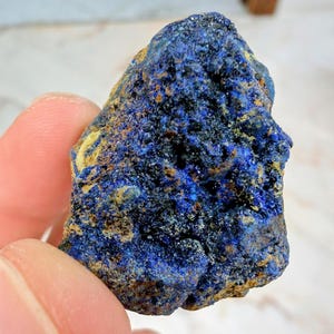 May include: A rough, natural azurite specimen. The stone displays a deep blue color with areas of lighter blue and hints of yellow and brown. The texture is uneven and crystalline, showcasing the mineral's natural formation.