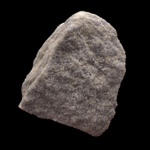 May include: A close-up of a gray rock against a black background. The rock has a rough, textured surface with various shades of gray and white. The shape is irregular, with a slightly rounded top and a jagged edge.
