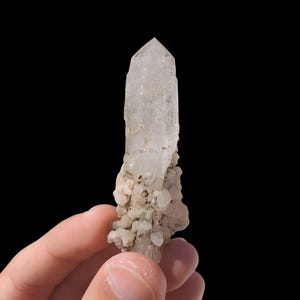 May include: A clear quartz crystal with a pointed tip, held against a black background. The crystal has a rough, textured surface and is partially covered with small, light-colored mineral deposits. The crystal is approximately 3 inches in length.