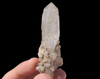May include: A clear quartz crystal with a pointed tip, held against a black background. The crystal has a rough, textured surface and is partially covered with small, light-colored mineral deposits. The crystal is approximately 3 inches in length.