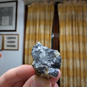 May include: A hand holding a small, irregularly shaped rock. The rock has a dark gray, metallic appearance with lighter gray and beige patches. The surface reflects light, suggesting a crystalline structure. The background includes framed artwork and yellow curtains.