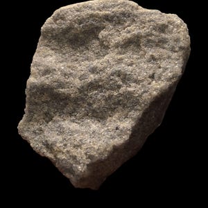 May include: A close-up of a light gray rock with a rough, textured surface. The rock has a slightly irregular shape and appears to be composed of small, granular particles. The background is solid black.