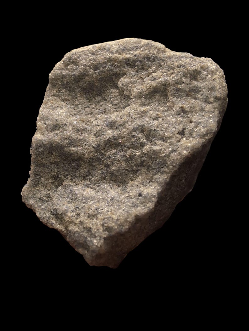 May include: A close-up of a light gray rock with a rough, textured surface. The rock has a slightly irregular shape and appears to be composed of small, granular particles. The background is solid black.