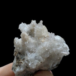 May include: A cluster of clear, crystalline quartz formations. The quartz crystals are small and needle-like, with a rough, textured appearance. The base of the cluster is a mix of gray and brown rock. The background is black.