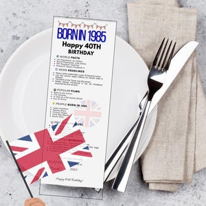May include: A card with the text "BORN IN 1985 Happy 40th Birthday" and facts from 1985, on a white plate with silverware and a linen napkin. A hand holds a UK flag. The card is a UK specific version.