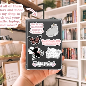 Może przedstawiać: Przezroczyste etui z naklejkami na Kindle. Naklejki obejmują motyla, kota, grzyba i tekst z napisem "this is my emotional support kindle" i "Treat People With Kindness". Obraz zawiera r&oacute;wnież tekst z napisem "Find all of these stickers and more on my shop to deck out your kindle, laptop, and more?"