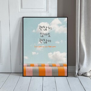 May include: A framed art print featuring a sky blue background with white clouds. The print has Korean text and the English phrase "It's okay to not feel okay." Below the sky is a row of orange, pink, and gray stripes.