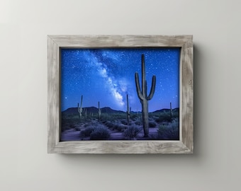 May include: A framed print featuring a desert landscape under a starry night sky. The image showcases tall saguaro cacti silhouetted against a deep blue sky filled with the Milky Way. The frame is made of weathered, light-colored wood.