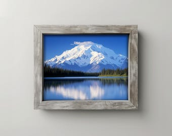 May include: A framed landscape photograph featuring a snow-capped mountain reflected in a calm lake. The image has a blue sky and a forest along the lake's edge. The frame is made of weathered wood.