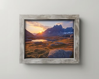 May include: A framed landscape print featuring a mountain range at sunset. The sky is filled with orange and yellow hues, with the sun setting behind the mountains. The foreground shows a small lake and rocky terrain.