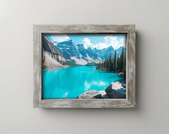 May include: A framed landscape print featuring a vibrant turquoise lake surrounded by snow-capped mountains and evergreen trees. The print is set within a rustic, weathered gray wooden frame, creating a natural aesthetic.