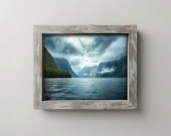 May include: A framed landscape photograph of a serene lake surrounded by lush green mountains under a cloudy sky. The frame is made of weathered, light-colored wood. The water is a deep blue, reflecting the overcast sky.