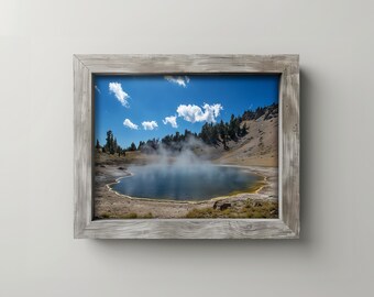 May include: A framed landscape photograph showing a lake with steam rising from the water. The sky is blue with white clouds. The frame is made of weathered wood.