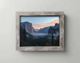 May include: A framed landscape photograph of a mountain valley at dawn. The image features a view of towering rock formations, a forest, and a layer of fog. The frame is made of weathered, light-colored wood.