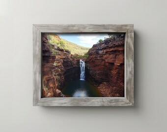 May include: A framed landscape photograph of a waterfall cascading into a pool of water. The image is set within a rustic, gray wooden frame. The canyon walls are reddish-brown, with green vegetation and a blue sky.