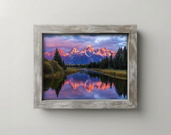 May include: A framed landscape print featuring a mountain range with snow-capped peaks reflecting in a calm river. The sky is filled with pink and purple hues, and the frame is a weathered gray.