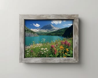 May include: A framed landscape photograph featuring a mountain range, a turquoise lake, and a vibrant field of wildflowers. The wooden frame has a weathered, gray finish. The sky is blue with scattered clouds.