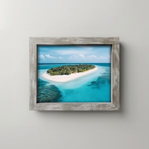 May include: A framed print featuring a tropical island with white sand, lush green vegetation, and turquoise water. The frame is made of weathered gray wood, adding a rustic touch to the serene landscape. The image captures a sunny day with a clear blue sky.