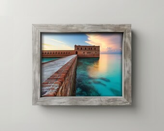 May include: A framed print featuring a scenic view of a brick pathway leading to a fort over turquoise water. The sky displays a colorful sunset with shades of pink, orange, and blue. The frame is made of weathered gray wood.