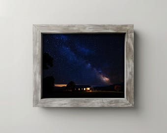 May include: A framed print featuring a night sky filled with stars and the Milky Way. The image shows a house with lit windows in the distance. The frame is made of weathered, light-colored wood.