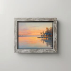 May include: A framed landscape painting depicts a serene lake scene at sunset. The sky transitions from orange to yellow, reflecting on the water. A cabin with lit windows sits on the shore, framed by trees. The frame is a weathered gray.