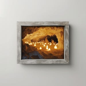 Op de afbeelding: Een ingelijste foto van een grotinterieur verlicht door talrijke kleine, gloeiende lichten. De grotwanden en het plafond hebben een warme, aardse tint, waarbij de lichten een zachte, uitnodigende sfeer creëren. De lijst is van verweerd, lichtgrijs hout.