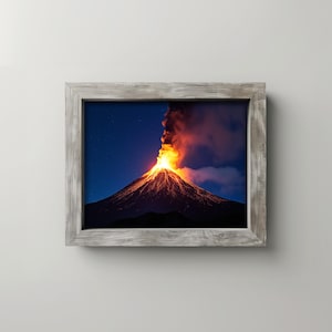 May include: A framed print of a volcanic eruption at night. The volcano is spewing bright orange lava and smoke against a dark blue sky with visible stars. The frame is made of weathered gray wood.