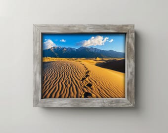May include: A framed landscape photograph of a desert scene. The image features a sandy dune with footprints leading towards a mountain range under a blue sky with clouds. The frame is made of weathered wood.
