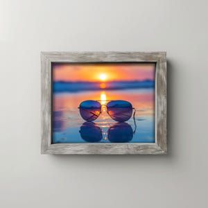 Puede incluir: Fotografía enmarcada de gafas de sol de aviador que reflejan una puesta de sol sobre el océano. Las gafas son negras con lentes azules, apoyadas sobre una superficie mojada. El marco es de madera gris envejecida.