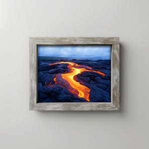 May include: A framed print featuring a vibrant image of flowing lava. The molten rock glows with shades of orange and yellow against a backdrop of dark, textured volcanic rock. The frame is a light gray color.