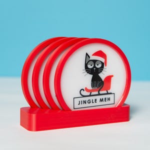 Puede incluir: Un juego de cuatro posavasos rojos y blancos con un soporte a juego. Cada posavasos presenta un gato negro con un gorro de Papá Noel y montado en un trineo, con el texto "JINGLE MEH". Los posavasos son redondos y brillantes.