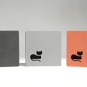 May include: Three square blocks in a row, each with a cat silhouette cutout. The blocks are dark gray, light gray, and orange. The cat design is black. The blocks are likely decorative or functional items.