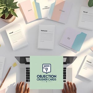 May include: An overhead shot of a workspace with a laptop, notebooks, and stationery. The notebooks have titles like "SALES" and "OBJECTIONS". The laptop screen displays the text "OBJECTION CRUSHER CARDS". A pink flower and a plant are also visible.