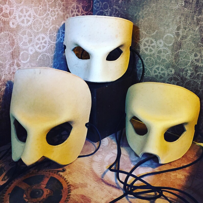 May include: Three white plaster masks with eye holes and a small mouth hole. The masks are on a dark surface with a background of a patterned wallpaper.