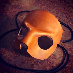 May include: A brown leather mask with eye and mouth cutouts. The mask has a black elastic strap for wearing.