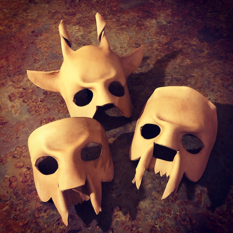 May include: Three beige leather masks with different facial features. The masks have eye holes and open mouths. The masks are on a brown textured surface.