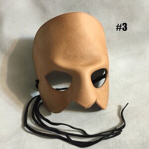 May include: A tan leather masquerade mask with eye holes and a jagged bottom edge. The mask has black leather ties.