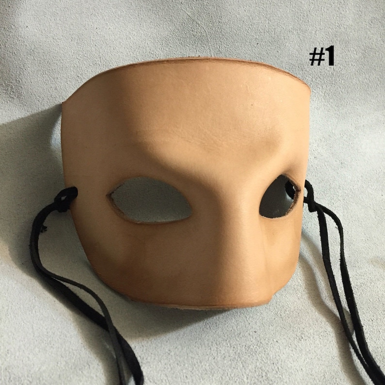 May include: A tan leather masquerade mask with black elastic ties. The mask has one eye hole and a raised, sculpted design.