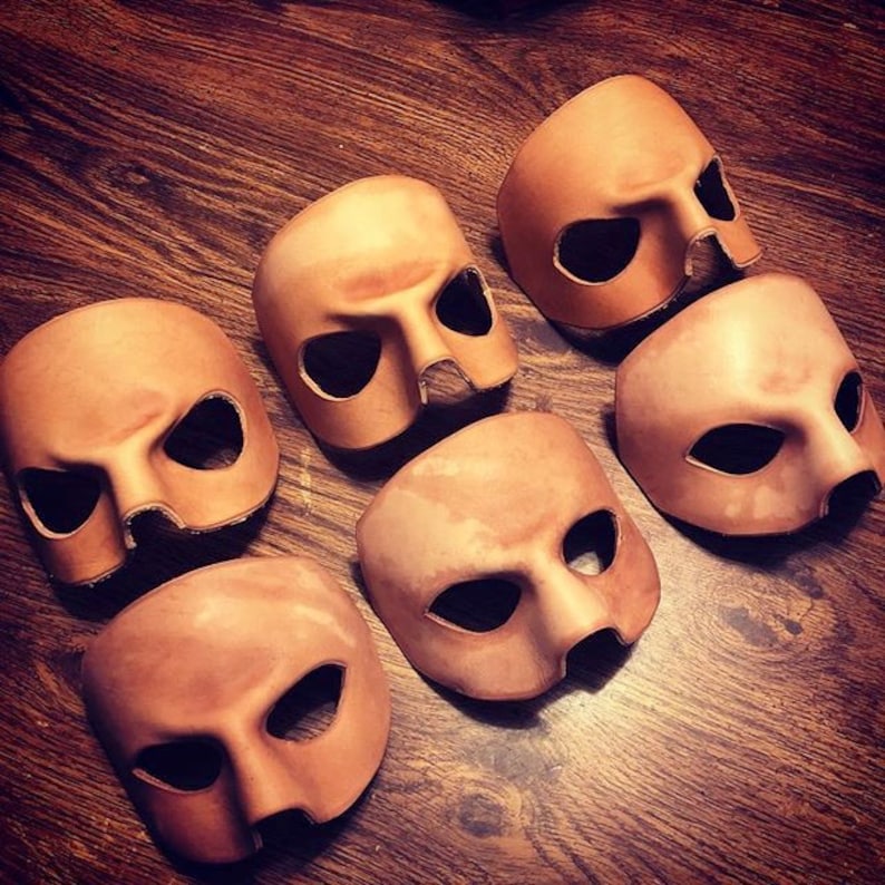 May include: Six brown leather masks with eye holes and a nose hole. The masks are arranged in a semi-circle on a wooden surface.