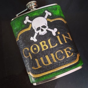May include: A stainless steel flask with a green and black leather cover. The cover features a white skull and crossbones and the words "GOBLIN JUICE" in gold lettering. The flask has a silver cap.