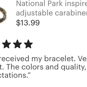 May include: A National Park-inspired adjustable carabiner bracelet. The bracelet is a braided design in shades of gray and green. The image also includes a customer review with five stars.