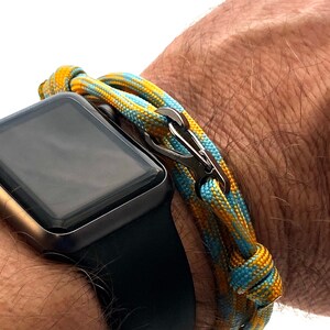 May include: A close-up of a wrist with a black smartwatch and a colorful bracelet. The bracelet is made of woven cord in shades of yellow, blue, and a silver clasp. The watch has a black band and a dark screen.